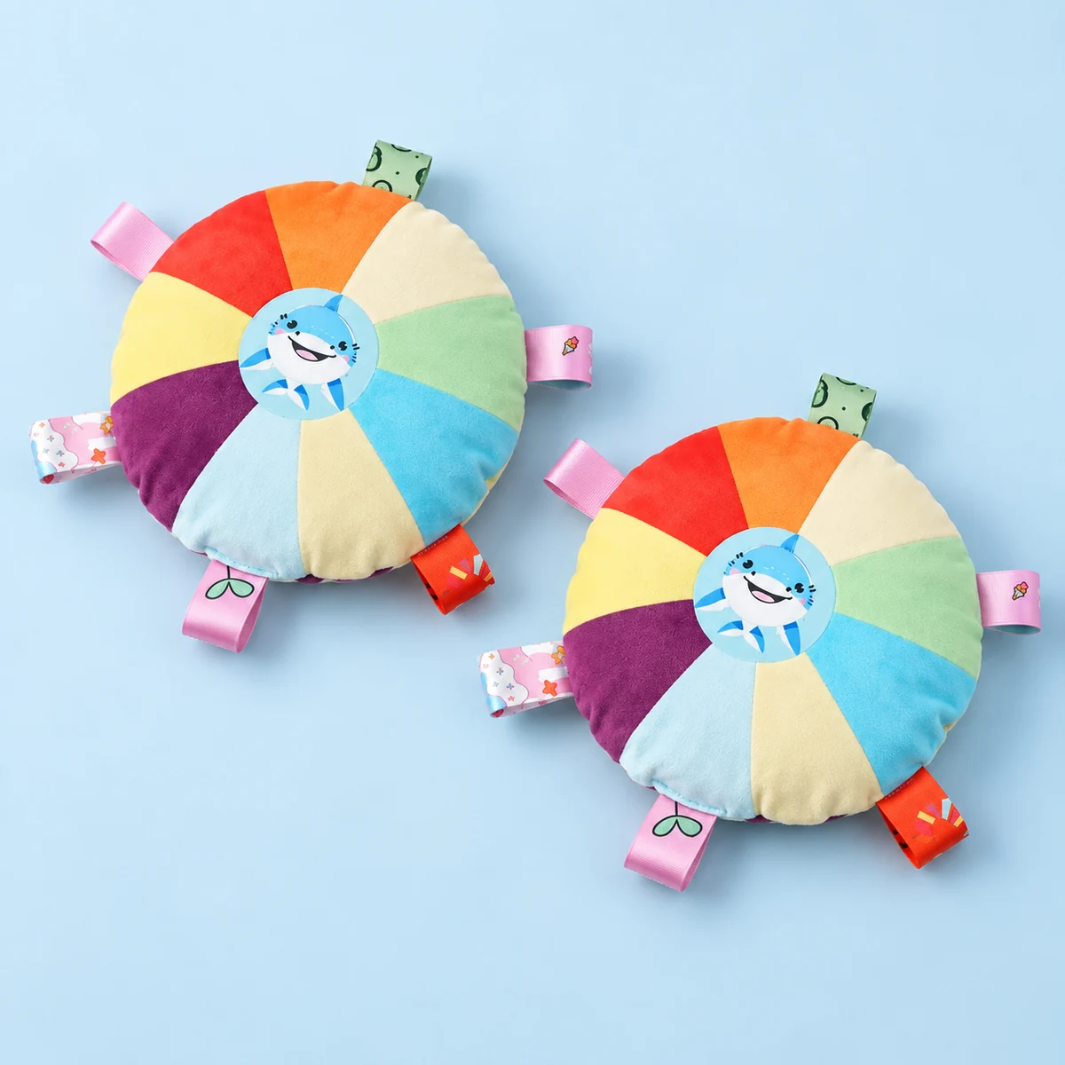 Two rainbow shark-print plush discs, each with six ribbon loops, on a pale blue surface.